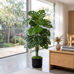 A large artificial monstera plant in a black pot, styled in a modern, sunlit living room next to a wooden coffee table.
