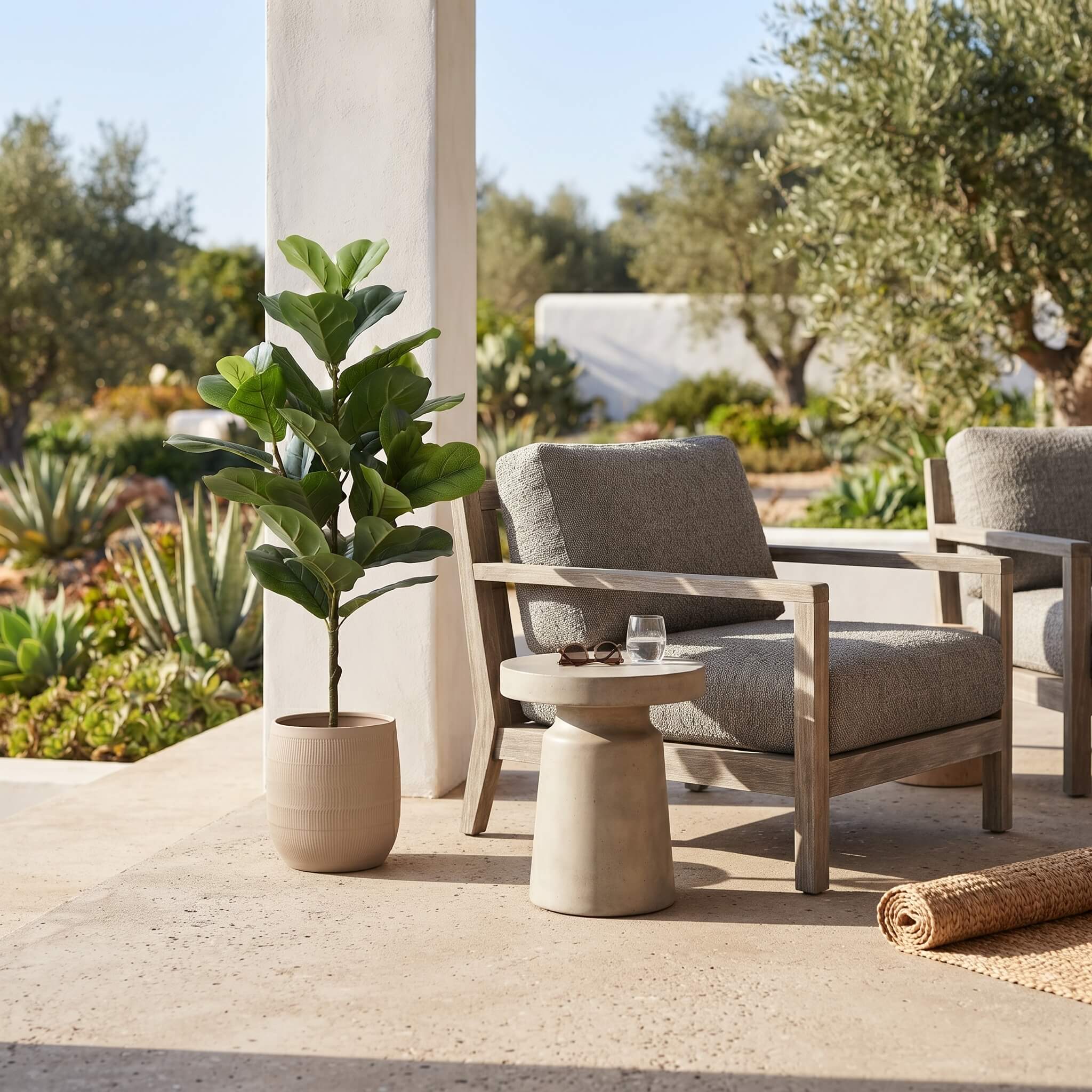 Outdoor patio furniture set with a light wood armchair, matching armchair partially visible, a round concrete side table, and a potted fiddle-leaf fig plant on a stone patio.