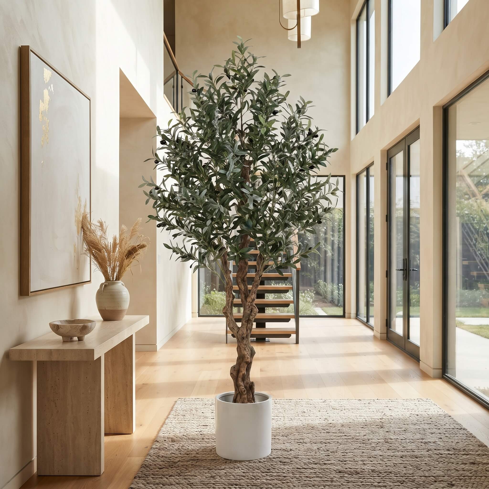 A tall potted olive tree with green leaves stands in a bright, modern interior hallway with wood floors, large windows, a console table, and a staircase in the background.