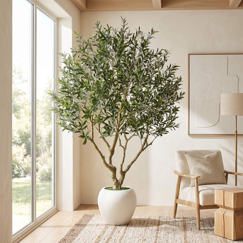 A tall artificial olive tree with green leaves and brown branches, potted in a white ceramic pot, placed in a bright, modern room next to a large window.
