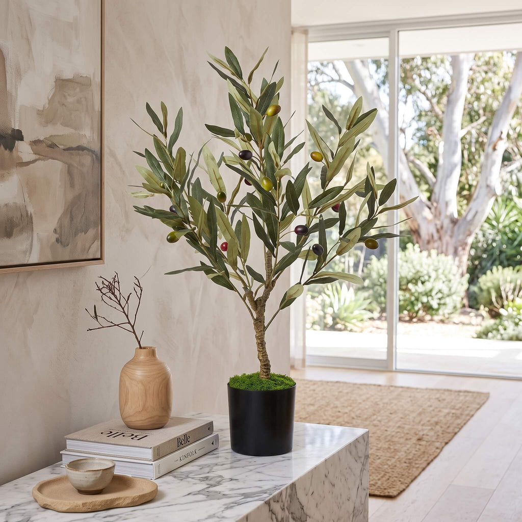An artificial olive tree in a black pot with faux greenery and small olives, displayed on a marble side table in a bright, modern room with a window and neutral decor.