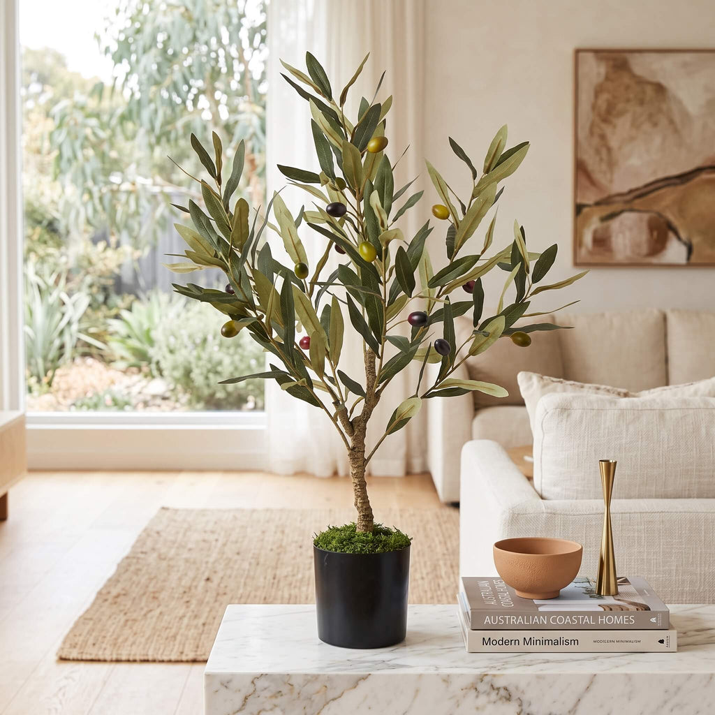 An artificial olive tree in a black pot with green and dark purple olives, styled on a marble coffee table in a bright, modern living room.