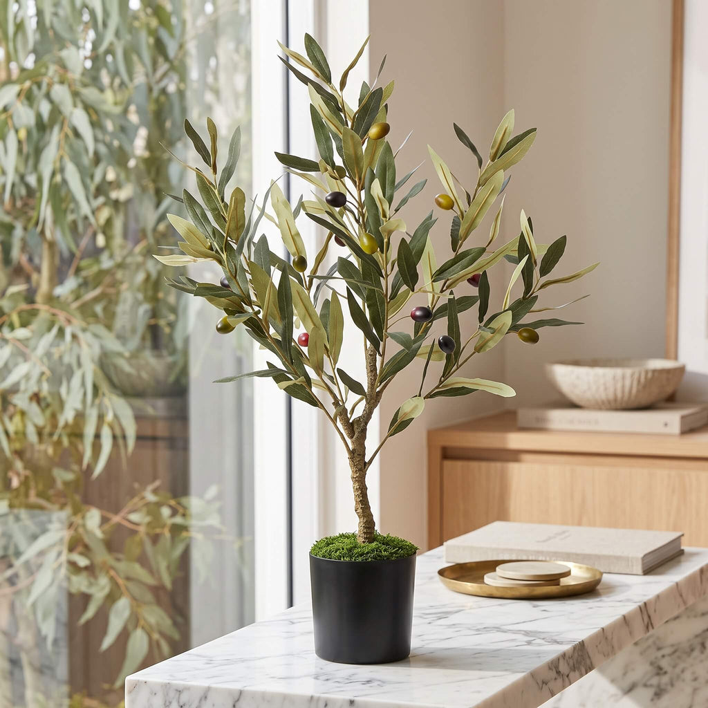 An artificial olive tree in a black pot with green and dark olives, displayed on a marble table in a bright, modern room.