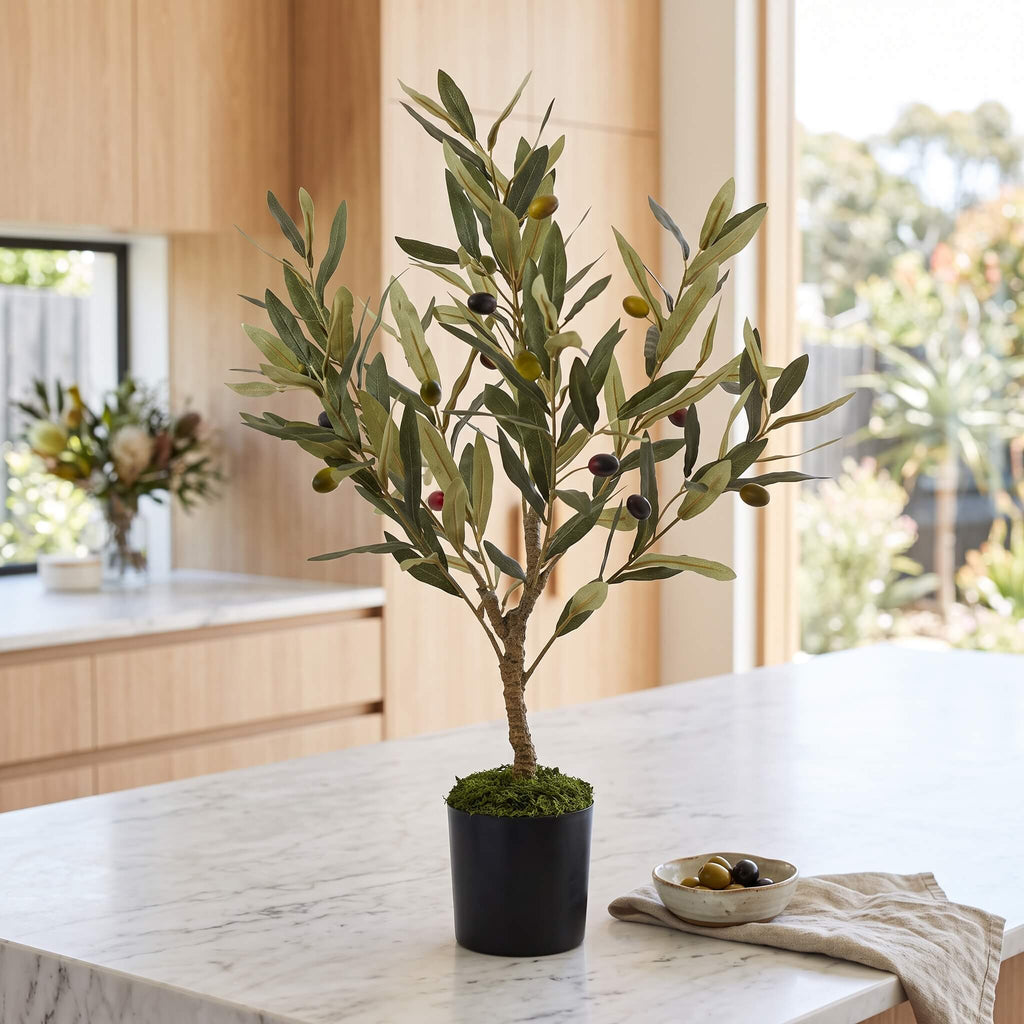 An artificial olive tree in a black pot with green and dark olives, displayed on a white marble kitchen island with a small bowl of olives nearby.