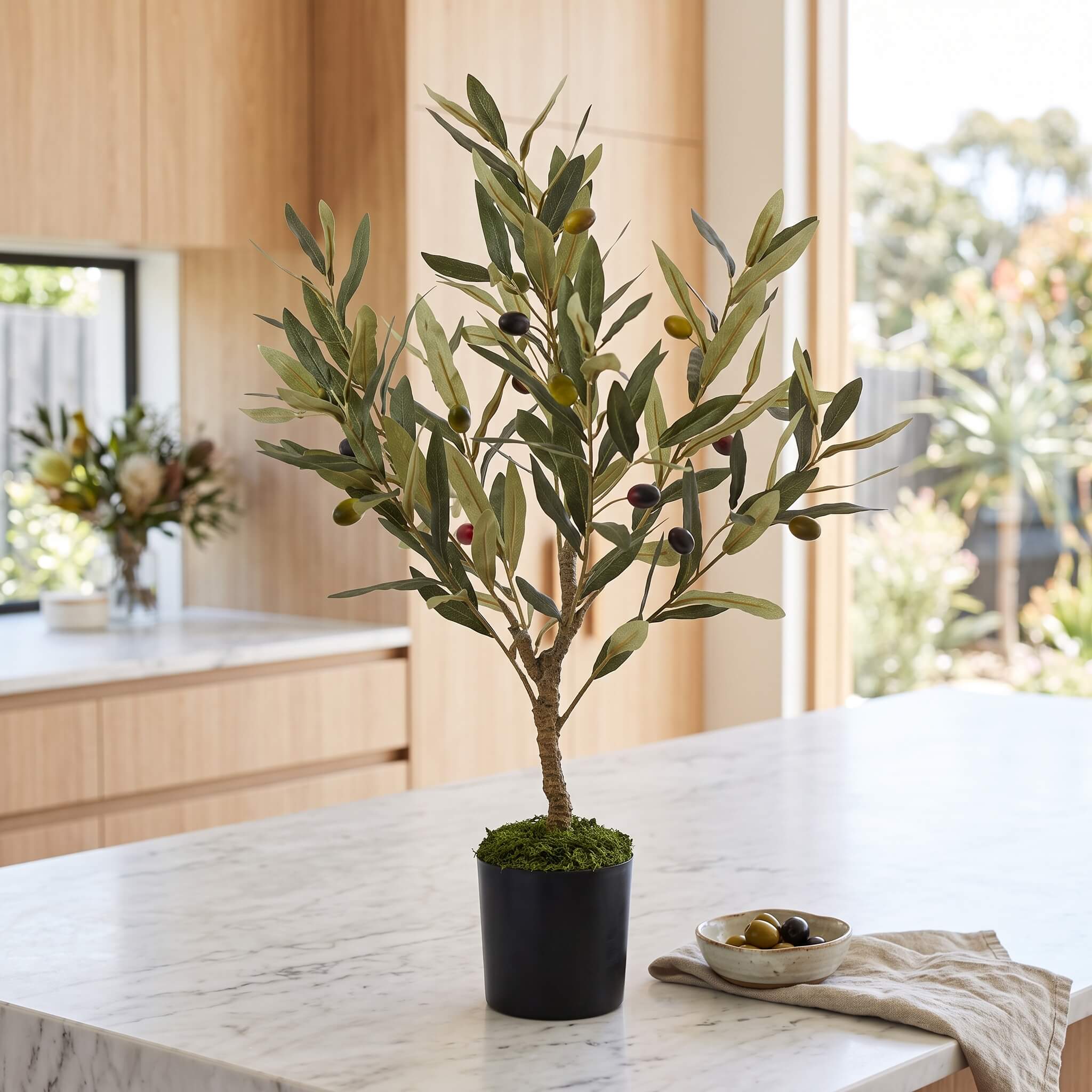 An artificial olive tree in a black pot with green and dark olives, displayed on a white marble kitchen island with a small bowl of olives nearby.