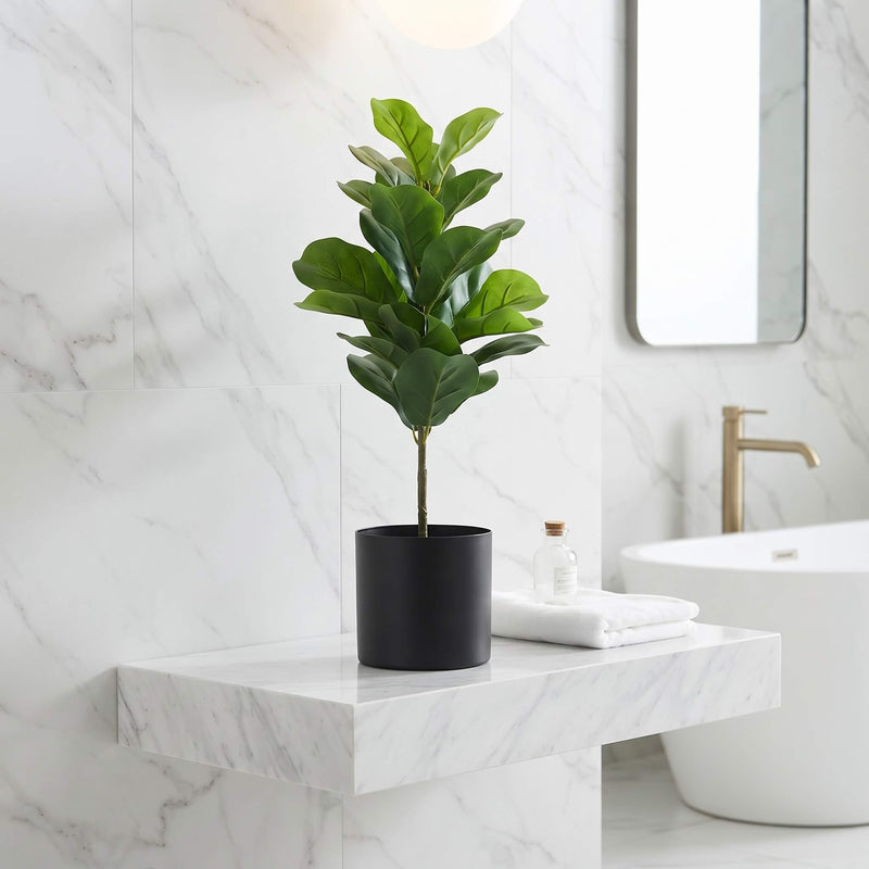 An artificial fiddle-leaf fig plant in a matte black cylindrical pot, styled on a white marble floating vanity in a modern bathroom setting.