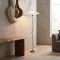 A modern floor lamp with a white pleated drum shade and a black wavy metal stem, standing in a minimalist living room next to a wooden bench and textured wall.