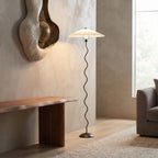 A modern floor lamp with a white pleated drum shade and a black wavy metal stem, standing in a minimalist living room next to a wooden bench and textured wall.