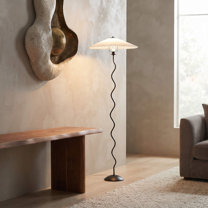 A modern floor lamp with a white pleated drum shade and a black wavy metal stem, standing in a minimalist living room next to a wooden bench and textured wall.