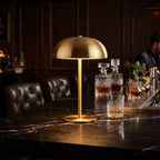 A modern table lamp with a brushed gold dome shade and matching gold stand sits on a dark marble bar counter in a dimly lit, upscale bar setting with leather chairs and a bartender in the background.