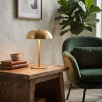 A modern brass dome table lamp sits on a rustic wooden side table in a styled living room setting next to a green velvet armchair and a potted fiddle-leaf fig plant.