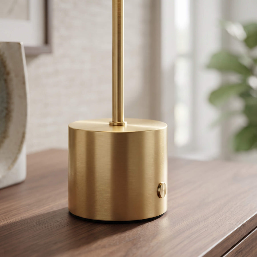 A modern cylindrical table lamp in brushed brass with a matching vertical stem, shown sitting on a dark wood table in a softly blurred indoor setting.