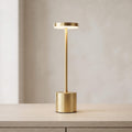 A modern table lamp with a brushed gold cylindrical base and stem, featuring a circular light panel on top that glows warm white, set on a light wood surface against a neutral beige background.