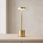 A modern table lamp with a brushed gold cylindrical base and stem, featuring a circular light panel on top that glows warm white, set on a light wood surface against a neutral beige background.