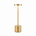 A modern minimalist floor lamp with a brushed gold finish, featuring a cylindrical base, a slim central stem, and a circular light-emitting top, shown against a plain white background.