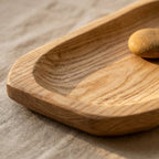 A close-up of a shallow wooden tray with a rounded, scalloped edge and visible wood grain, resting on a textured beige fabric surface with a small smooth stone inside.
