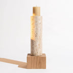 A modern cylindrical candle holder made of speckled travertine stone, illuminated from within to cast a warm glow, with a beige pillar candle on top and a light wood block base.