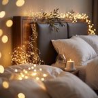 A cozy bedroom scene with a bed dressed in white bedding, a dark upholstered headboard, warm string lights draped around the headboard and across the room, a lit candle on a bedside table, and a glass carafe.