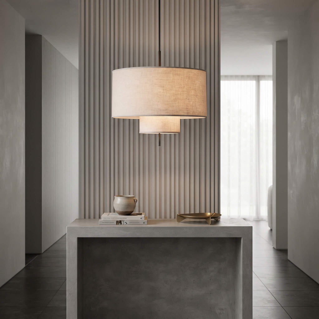 A modern two-tier drum pendant light with a beige linen shade hangs over a gray concrete console table in a minimalist, contemporary interior hallway.