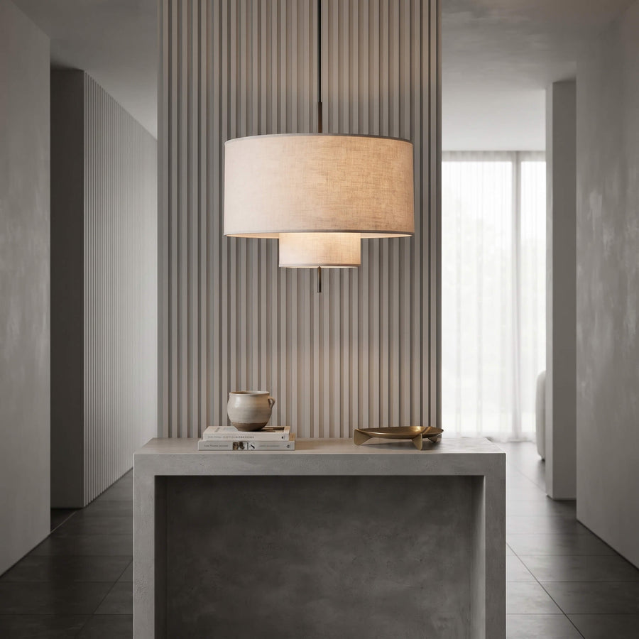 A modern two-tier drum pendant light with a beige linen shade hangs over a gray concrete console table in a minimalist, contemporary interior hallway.