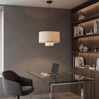 A modern two-tier drum pendant light with a beige fabric shade hangs over a glass-top desk in a contemporary home office setting with a gray chair and built-in wooden shelves.