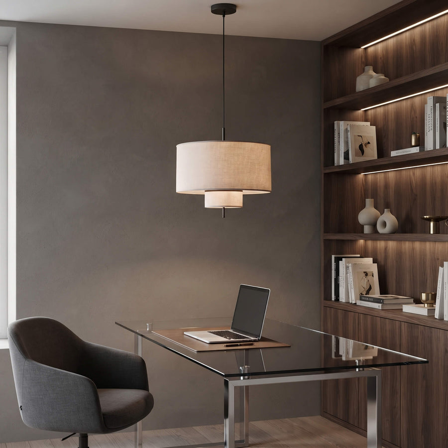 A modern two-tier drum pendant light with a beige fabric shade hangs over a glass-top desk in a contemporary home office setting with a gray chair and built-in wooden shelves.