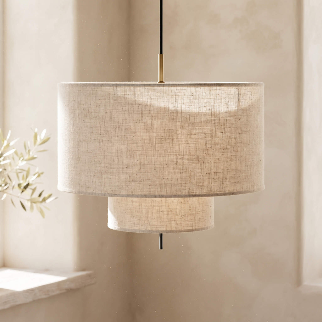 A modern drum pendant light with a textured beige linen shade, featuring a two-tiered design with a larger top drum and a smaller bottom cylinder, suspended by a black cord with a brass accent.