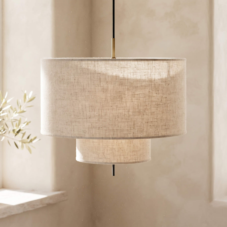 A modern drum pendant light with a textured beige linen shade, featuring a two-tiered design with a larger top drum and a smaller bottom cylinder, suspended by a black cord with a brass accent.