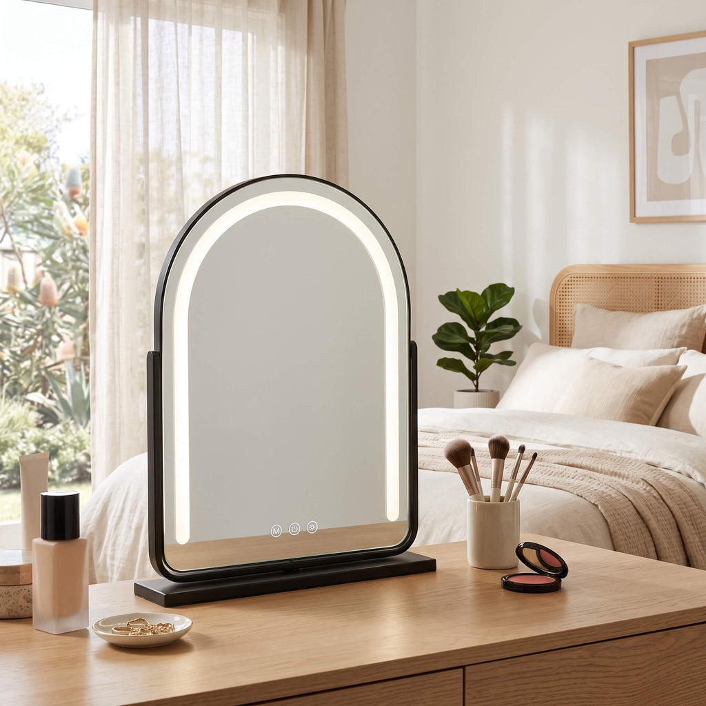 An arched LED vanity mirror with a black frame and base, illuminated around the mirror surface, set on a wooden dresser in a bright, modern bedroom.