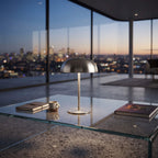 A modern dome-shaped table lamp with a brushed metal finish sits on a glass coffee table in a high-rise office or lounge, with a city skyline at dusk visible through the large windows behind it.