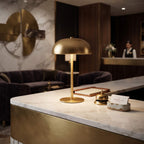 A modern dome-shaped table lamp in brushed gold brass sits on a marble-topped counter in a luxury hotel lobby. The background features dark wood paneling, velvet seating, and a reception desk with a waiter visible in the distance.
