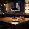 A modern black table lamp with a dome shade and slim stem sits on a wooden bar table in a dimly lit bar or lounge. A glass of amber liquor and a small plate with a lemon wedge are placed beside it, with blurred bar seating and shelves in the background.