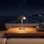 A modern dome-shaped table lamp with a brushed metal finish sits on a wooden dining table, casting a warm glow under a starry night sky and ocean backdrop.