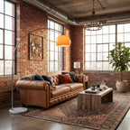A styled living room featuring a brown leather Chesterfield sofa with blue and rust accent pillows, a rustic wooden coffee table, a large patterned area rug, a silver floor lamp, and a warm orange dome pendant light against a brick wall background.