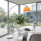 A modern arched floor lamp with a bright orange dome shade and silver metal frame stands beside a white dining table and chairs in a bright room with large windows and potted plants.