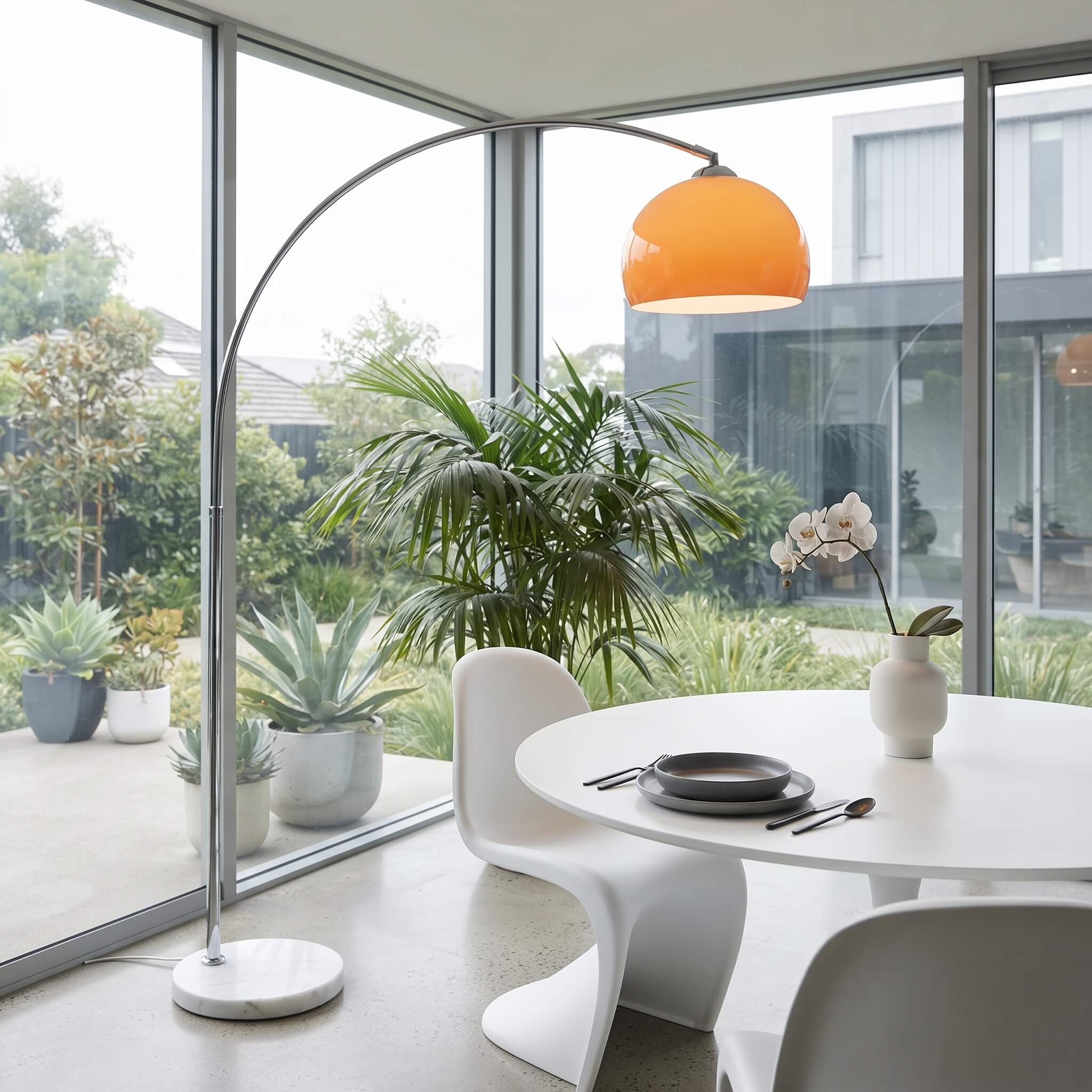 A modern arched floor lamp with a bright orange dome shade and silver metal frame stands beside a white dining table and chairs in a bright room with large windows and potted plants.