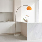 A modern arched floor lamp with a warm orange dome-shaped shade and a slim metallic stand, placed in a bright, minimalist kitchen with white cabinets and a marble island.
