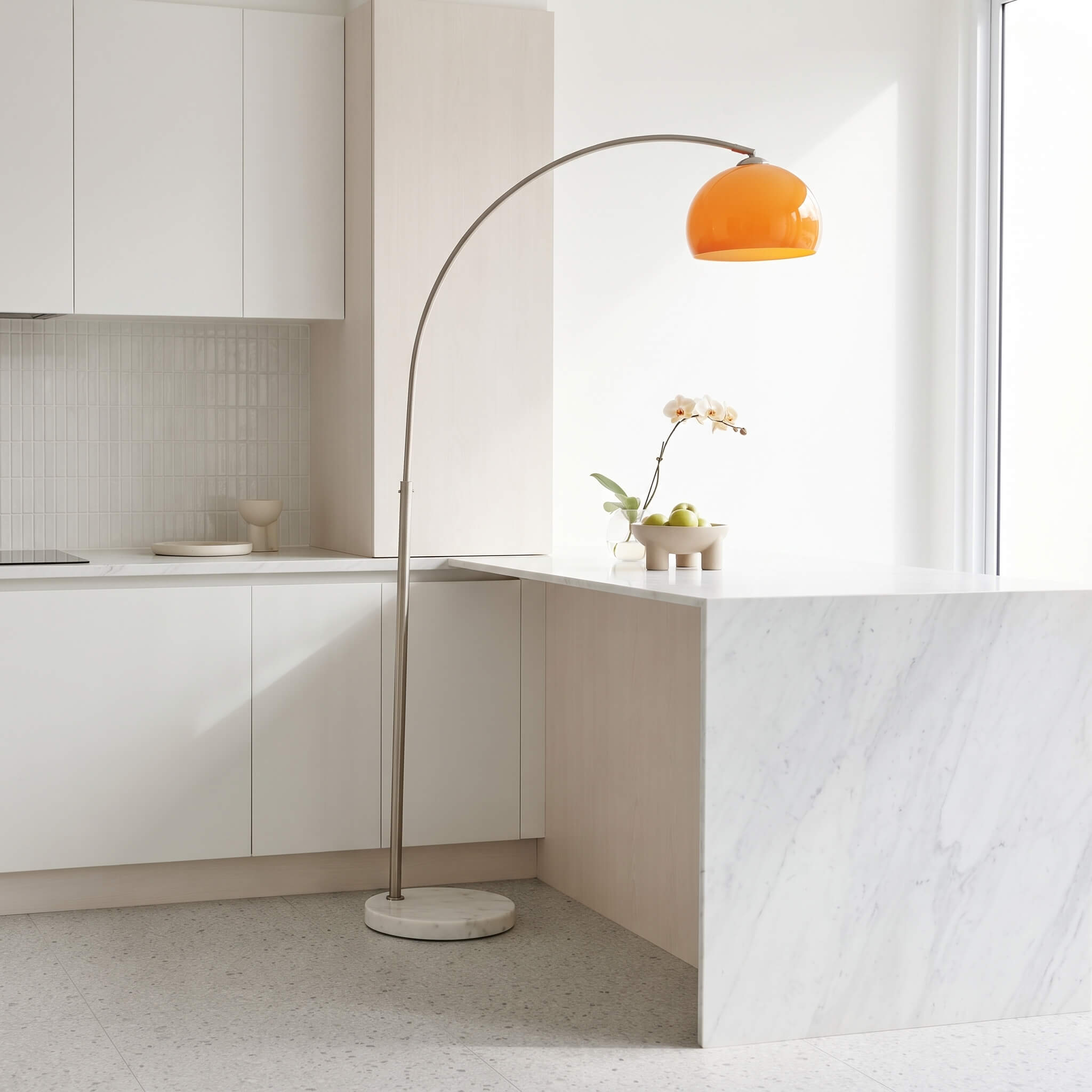 A modern arched floor lamp with a warm orange dome-shaped shade and a slim metallic stand, placed in a bright, minimalist kitchen with white cabinets and a marble island.