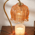 An amber-orange ribbed glass table lamp with a curved brass-colored metal arm, shown lit above a white candle on a wooden surface against a light wall.