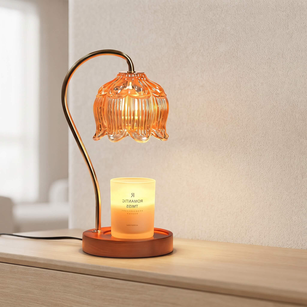 A lit amber glass flower-shaped table lamp with a curved metallic stem and a glowing candle holder in a round wooden base, placed on a light wood surface against a neutral wall.