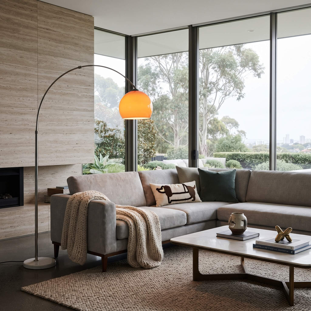 A modern living room featuring a light gray sectional sofa with decorative pillows and a knit throw blanket, next to a tall arched floor lamp with a glowing orange shade. The room has large windows with a view of trees, a beige stone wall, and a low wooden coffee table.