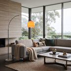 A modern living room featuring a light gray sectional sofa with decorative pillows and a knit throw blanket, next to a tall arched floor lamp with a glowing orange shade. The room has large windows with a view of trees, a beige stone wall, and a low wooden coffee table.