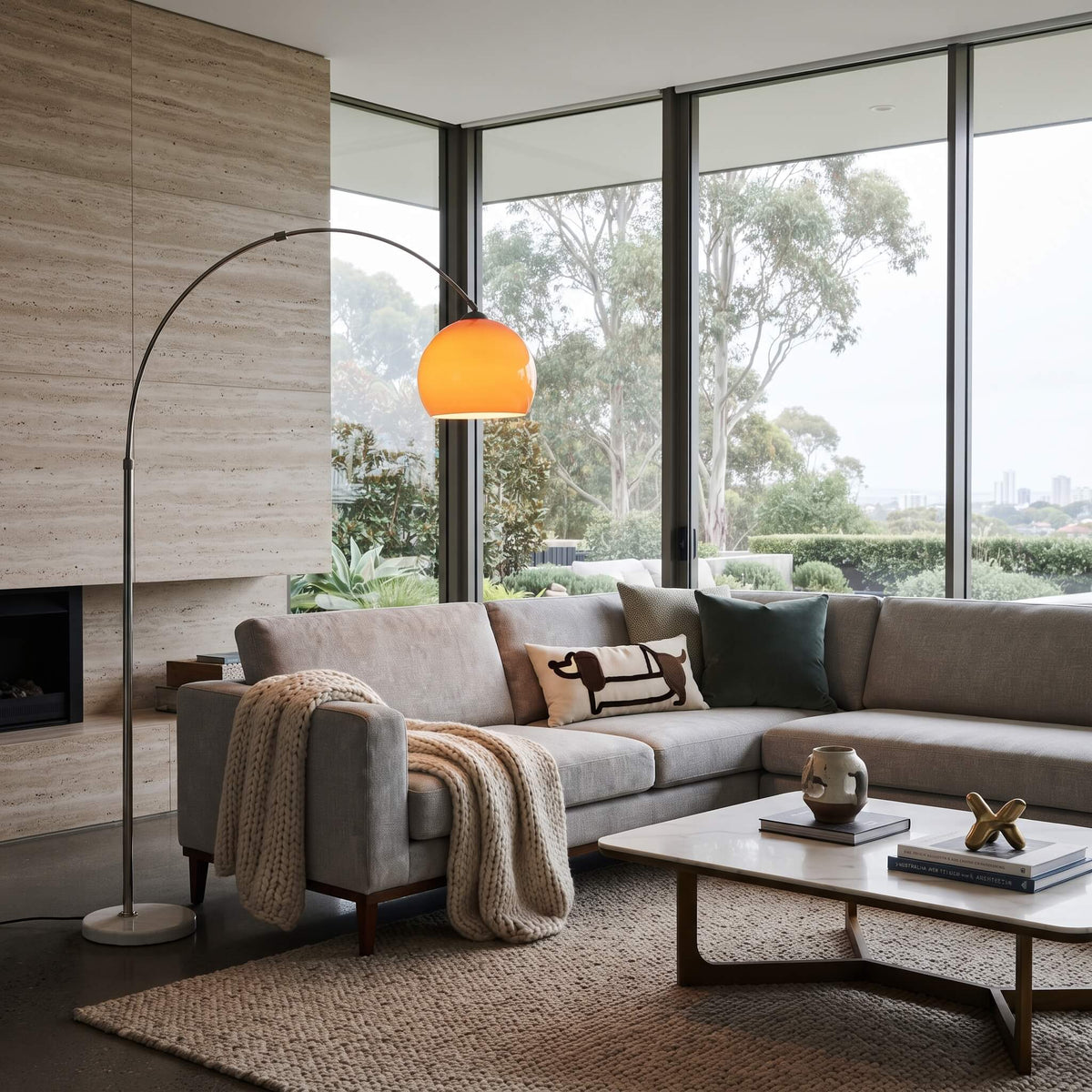 A modern living room featuring a light gray sectional sofa with decorative pillows and a knit throw blanket, next to a tall arched floor lamp with a glowing orange shade. The room has large windows with a view of trees, a beige stone wall, and a low wooden coffee table.