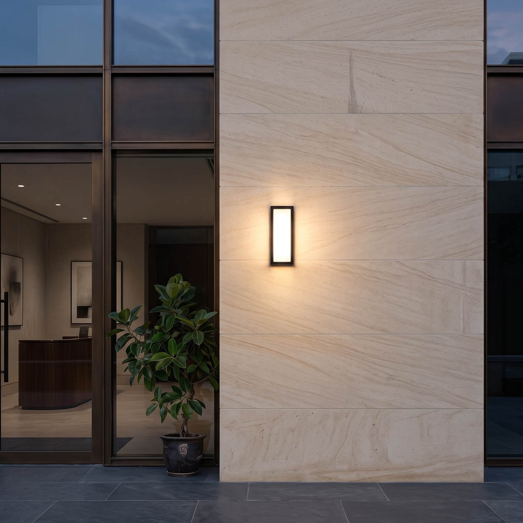 Sentry motion sensor wall light glowing warm on a sandstone wall beside a glass entry at dusk
