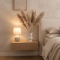 A warm, cozy bedroom scene featuring a light wood floating nightstand with a lit ceramic table lamp and a white vase filled with pampas grass, set against a textured beige wall next to a bed.