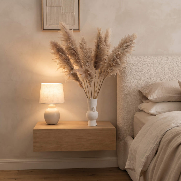 A warm, cozy bedroom scene featuring a light wood floating nightstand with a lit ceramic table lamp and a white vase filled with pampas grass, set against a textured beige wall next to a bed.