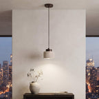 A modern cylindrical pendant light with a dark metal rod and a textured light gray shade hangs over a dark wood console table in a minimalist room. Large windows in the background show a city skyline at dusk.