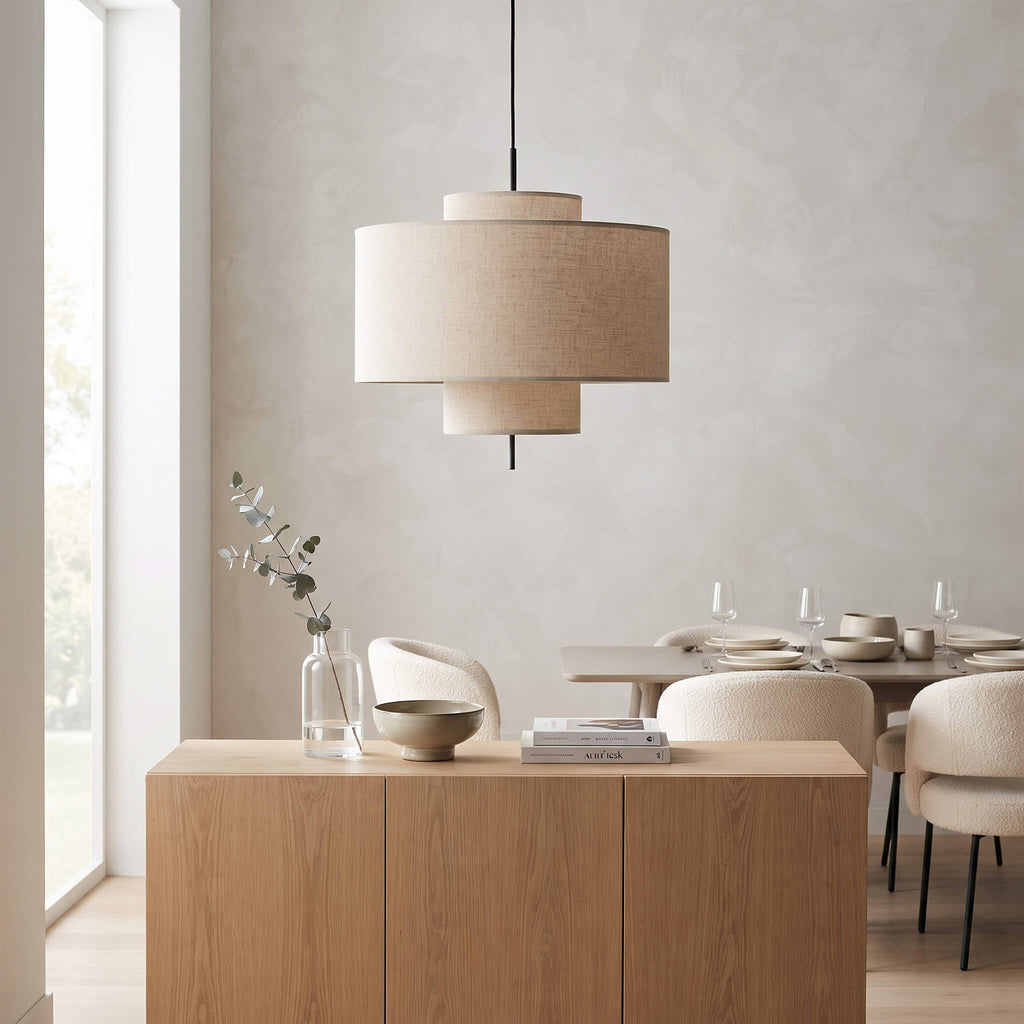A modern two-tiered drum pendant light with beige linen shades hangs over a light wood sideboard in a minimalist dining room setting.