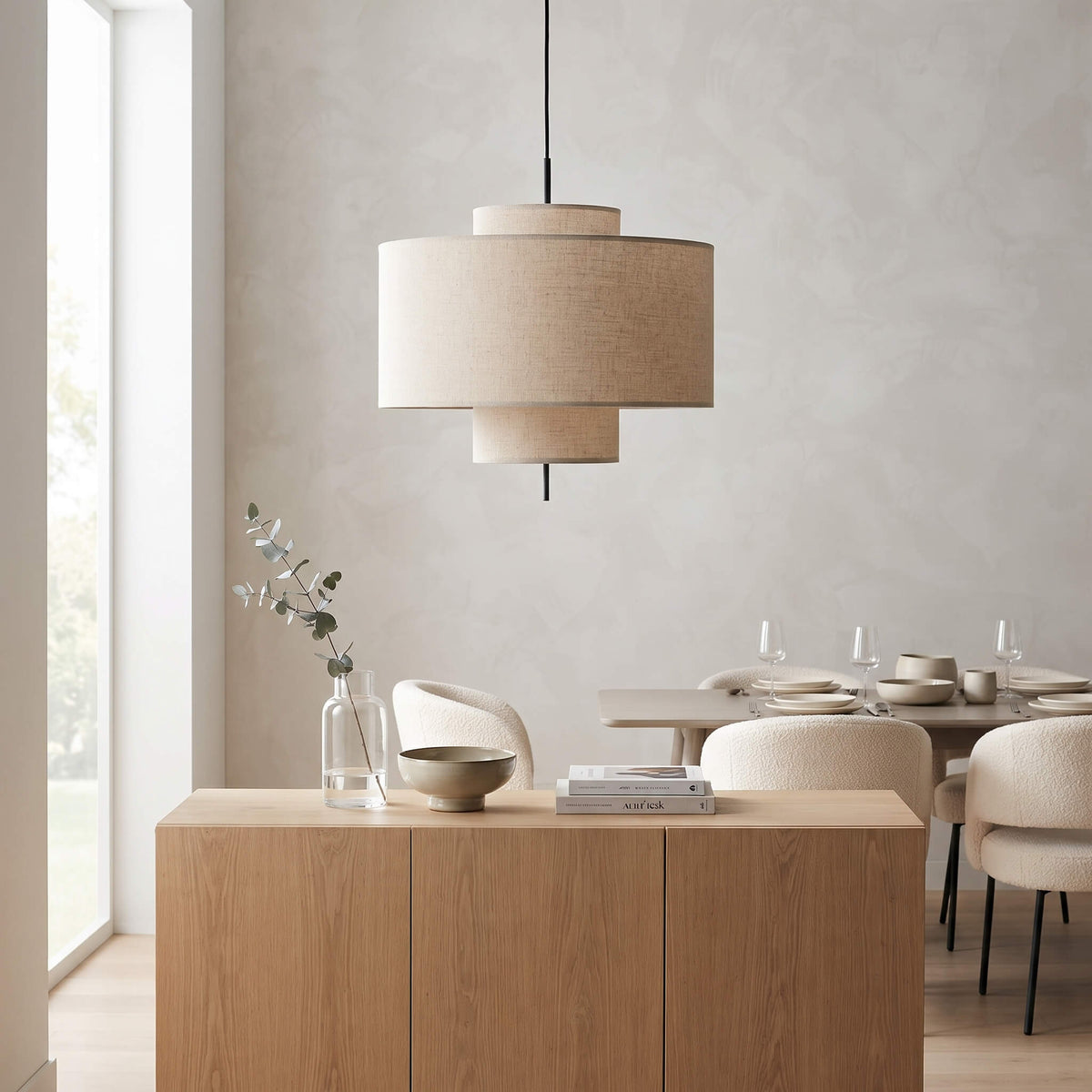 A modern two-tiered drum pendant light with beige linen shades hangs over a light wood sideboard in a minimalist dining room setting.