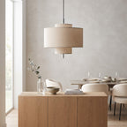 A modern two-tiered drum pendant light with beige linen shades hangs over a light wood sideboard in a minimalist dining room setting.